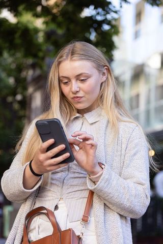 Adult woman scrolling smartphone on city sidewalk in cardigan with leather crossbody bag