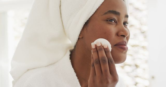 Image of happy african american woman in robe cleansing face. beauty treatment and skin care routine concept.
