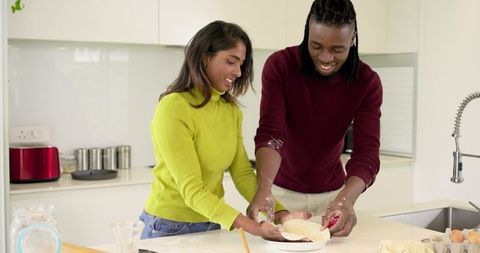 Interracial couple baking and kneading dough on kitchen island wearing colorful sweaters
