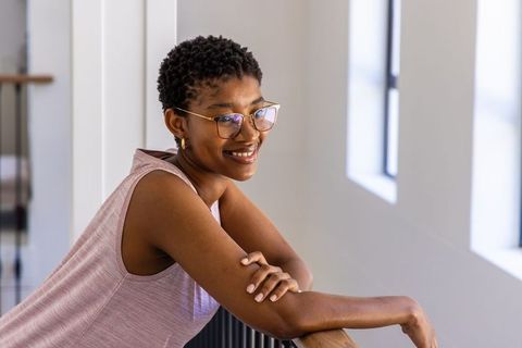 Smiling african woman leaning on railing in bright minimalist hallway wearing gold glasses