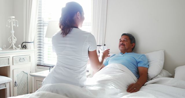Caregiver assisting an elderly man in bed by providing a glass of water. Ideal for use in healthcare and nursing-related content, retirement and aged care promotions, patient recovery guides, and home care assistance marketing.