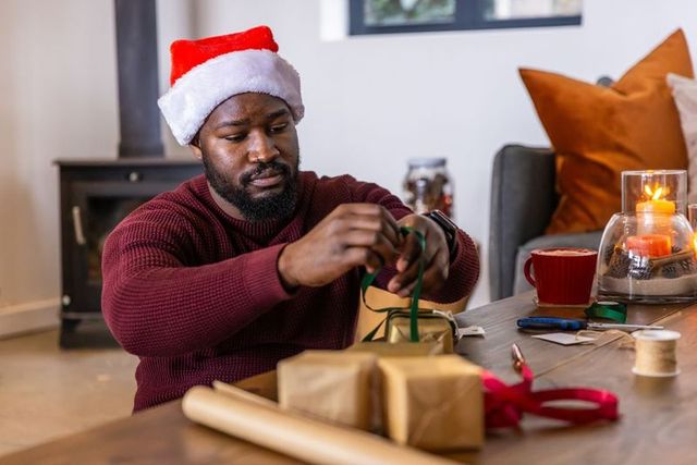 African american man wearing santa hat tying green ribbon on kraft-wrapped christmas gift