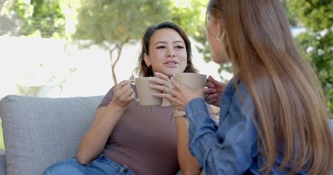 Two women sitting on outdoor sofa holding coffee mugs and talking in relaxed sunny backyard
