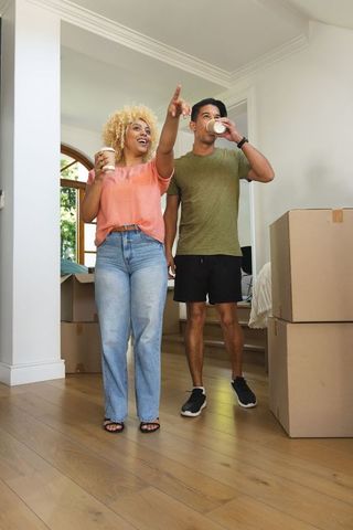 Diverse couple unpacking boxes in bright home, pointing at new bedroom with hardwood floors