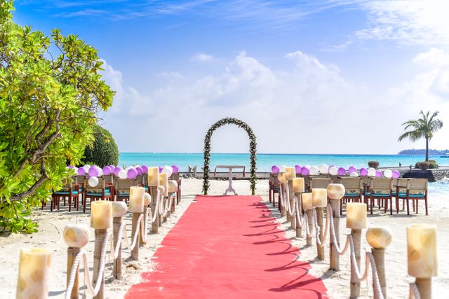 This image depicts a stunning beach wedding setup by the ocean, featuring a red carpet leading to an archway decorated with flowers. Wooden chairs adorned with purple bows are lined up on either side of the carpet, with candles on stands completing the festive look. The clear blue sky, white sandy beach, and calm sea provide an idyllic setting. Ideal for use in wedding planning websites, travel blogs, or magazines promoting tropical and destination weddings.