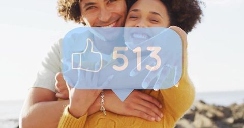 Sunlit embracing couple wearing mustard sweater on rocky beach with social like notification bubble