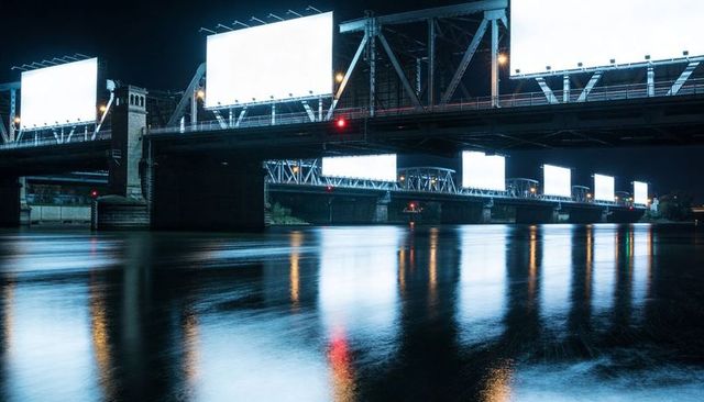 Nighttime steel truss bridge lined with illuminated blank billboards reflecting on river