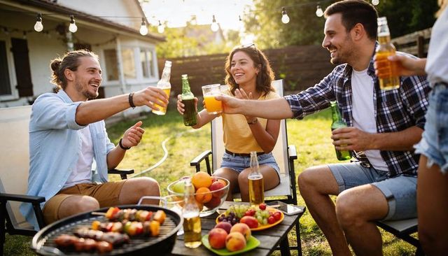 Young friends toasting at backyard bbq celebrating summer with grill, drinks and string lights