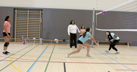 Diverse women lunging for volleyball on indoor court during team practice and coaching