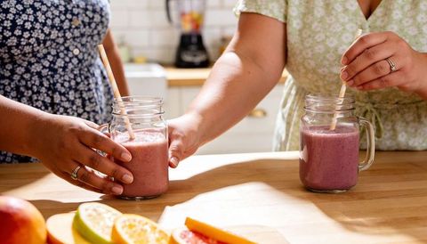 Two women stirring pink berry smoothies in mason jars on bright kitchen counter