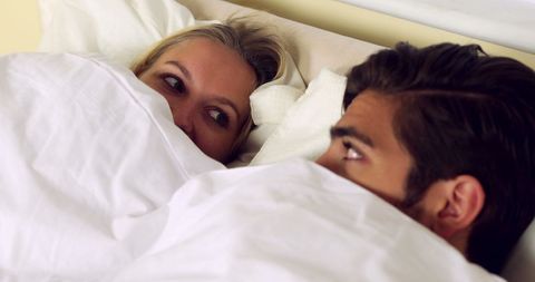 Playful couple hiding beneath sheets at home in bedroom