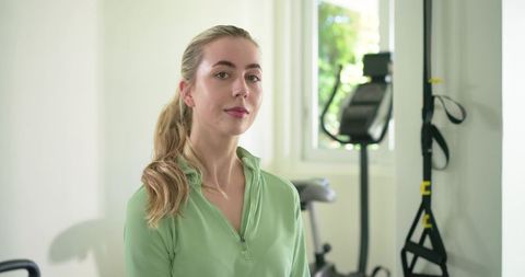 Young woman standing in home gym wearing green zip-up, trx straps and exercise bike