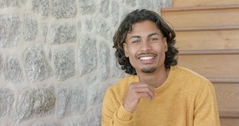 Young man sitting on wooden staircase smiling in mustard knit sweater against textured stone wall