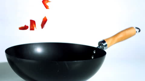 Sliced Red Pepper Falling into Black Wok Over White Background