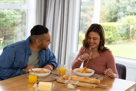 Diverse couple sharing spaghetti and baguette at sunlit dining table, smiling together