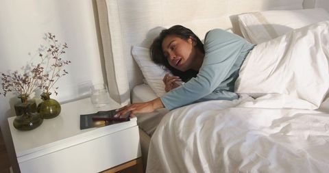 Asian woman reaching for phone while lying in cozy minimalist bed with soft morning light