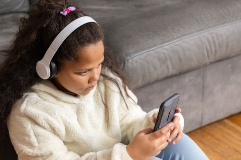 African american girl listening with headphones holding smartphone on living room floor