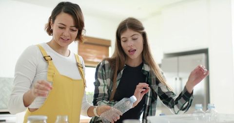 Roommates examining plastic bottles and glass jars in sunlit modern kitchen for recycling