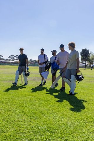 Five cricketers walking across sunny outfield carrying bats helmets and protective pads
