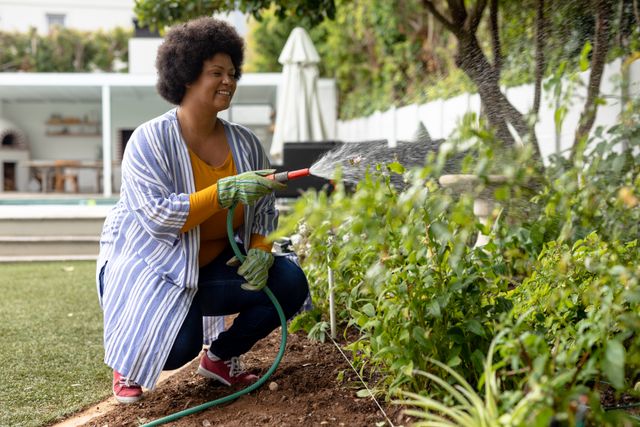Smiling african american mid adult woman watering plants in backyard garden. unaltered, lifestyle, leisure activity, gardening hose, protection and gardening concept.