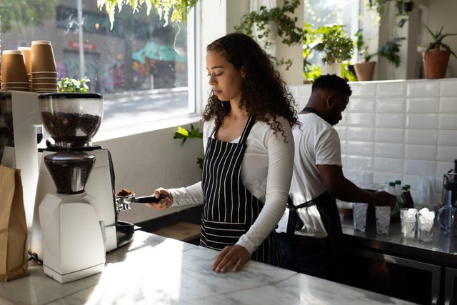 Biracial female barista using coffee maker against african american coworker in cafe kitchen. unaltered, cafe culture, people and occupation concept.
