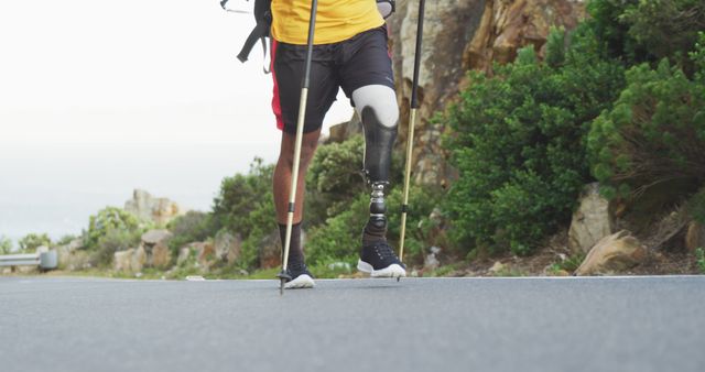 Low angle view of fit, disabled biracial man with prosthetic leg, enjoying his time on a trip to the mountains, hiking, walking on the road, using Nordic walking sticks, on a cloudy day