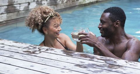 Couple toasting in pool by weathered wooden deck, sharing cocktail smiles