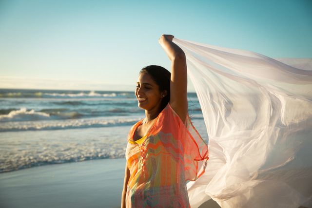 Smiling biracial woman having fun, dancing on beach holding scarf. holiday, freedom and leisure time at the beach.