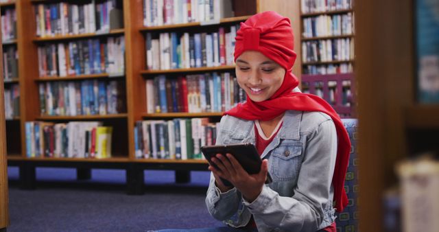 Young girl with red hijab using tablet seated against library bookshelves. Ideal for educational content, digital learning, multicultural themes, student resources, e-learning, and promoting reading habits in modern environments.