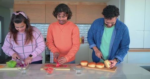 Diverse friends preparing burgers at kitchen island, slicing tomatoes, onions and burger buns