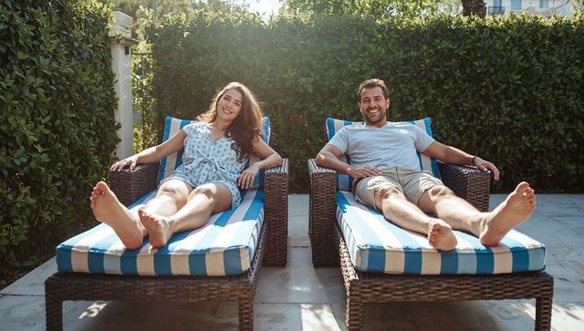 Smiling couple relaxing on striped wicker chaise lounges in sunlit backyard patio
