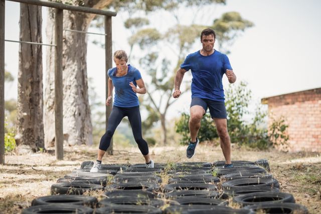 Man and woman running over tires in an outdoor boot camp obstacle course. They are focused and determined, showcasing teamwork and physical fitness. Ideal for use in fitness blogs, training programs, motivational posters, and advertisements for outdoor activities or fitness events.