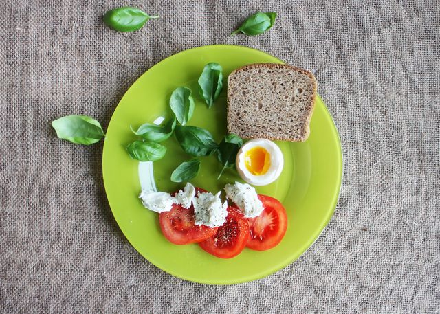 Colorful breakfast plate featuring soft-boiled egg, whole grain bread slice, tomato slices, and fresh basil leaves, arranged beautifully on green plate. Ideal for articles or promotions about healthy eating, vegetarian diets, and nutritious meal ideas. Great for food blogs, restaurant menus, and health-focused content.