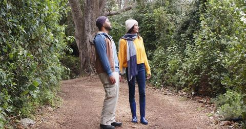 Diverse couple exploring wooded trail in beanies and scarves, gazing up at canopy
