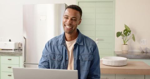 Young Man in Denim Shirt Smiling While Working at Laptop in Bright Kitchen