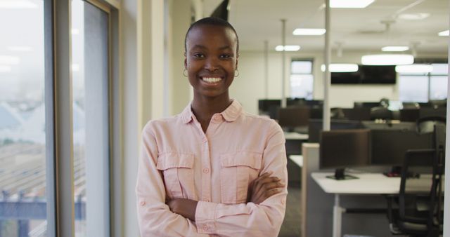 African American woman wearing glasses standing confidently with arms crossed in an open office. Ideal for usage in business and corporate content, showcasing diversity, leadership, confidence, and workplace scenarios.