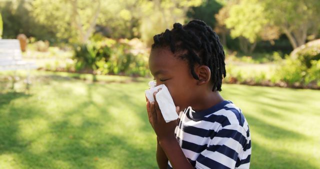A young African American boy is blowing his nose with a tissue outdoors, with copy space. Seasonal allergies or a common cold might be causing discomfort for the child in a garden setting.