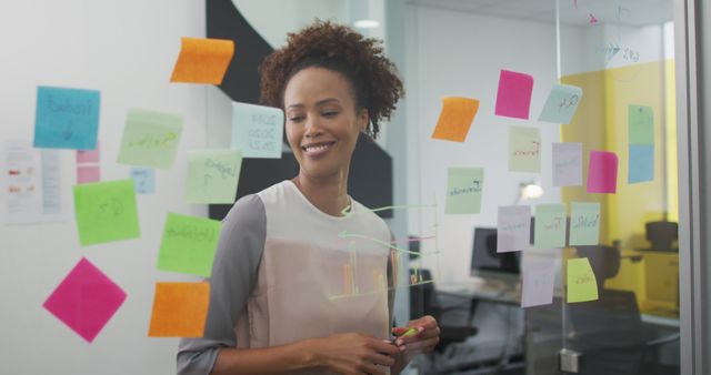 Smiling biracial woman standing by transparent board with memo notes in office. business professional and working in busy modern office.
