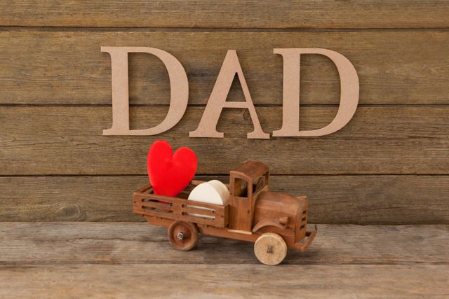 Wooden toy truck carrying red and white heart shapes in front of 'Dad' text on rustic wooden table. Ideal for Father's Day cards, family-themed decorations, and gifts celebrating fatherhood.