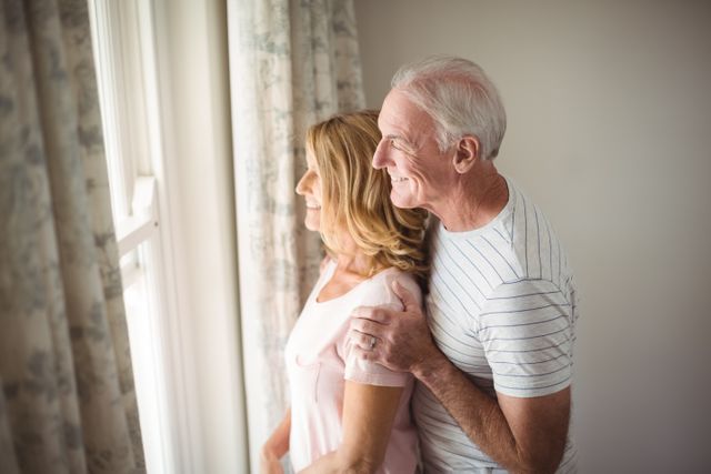 Senior couple enjoying moment looking out window, suggesting happiness and togetherness. Suitable for themes of love, aging, domestic life, and health. Ideal for articles on relationships, retirement, home living, and well-being.