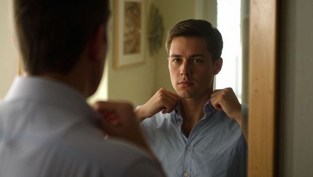 Young man adjusting light-blue button-up shirt collar in mirror preparing for day
