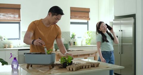Asian couple planting herbs on kitchen island, man using trowel, woman speaking on phone
