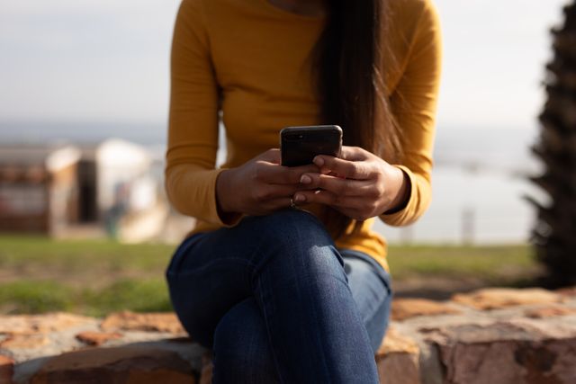 Woman sitting outdoors on a sunny day, using a smartphone. Ideal for themes related to technology, communication, leisure, and outdoor activities. Can be used in articles, blogs, and advertisements focusing on mobile technology, casual lifestyle, and relaxation.