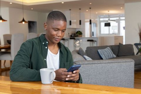 African american man checking smartphone while enjoying coffee in modern open living room