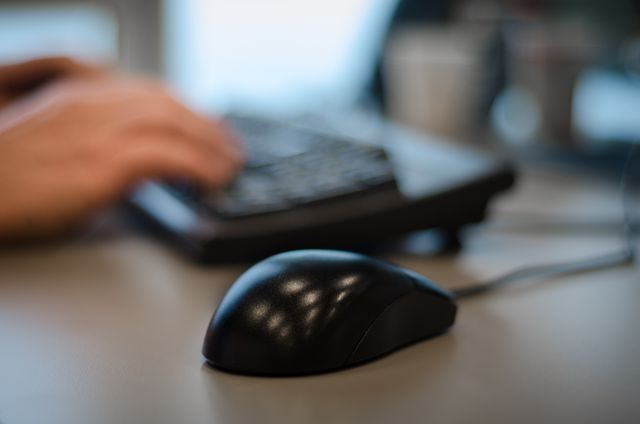 Close-up view of a computer mouse on a desk with a blurred background of a person's hand typing on a keyboard. Suitable for illustrating technology usage in the workplace, online work, office environments, or business-related themes.