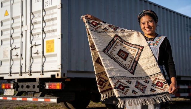 Smiling artisan woman holding handwoven rug with geometric design near shipping trailer