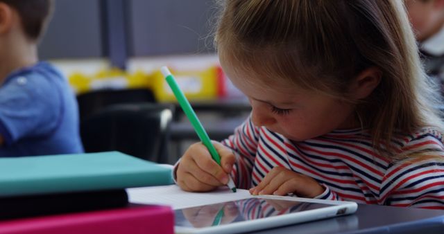 Young girl concentrating deeply on writing in a notebook at a classroom desk, surrounded by school supplies like books and a tablet. Useful for educational resources, school advertisements, teaching materials, and articles on childhood education.