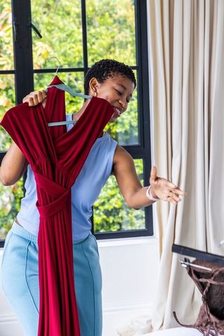 African woman packing maroon gown into open suitcase while smiling by sunlit window