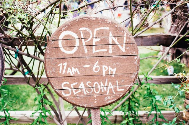 This rustic wooden sign, indicating open seasonal hours from 11 am to 6 pm, is ideal for businesses or attractions with specific seasonal timings. The hand-painted letters on the weathered wood offer a charming, vintage feel. Use this image to represent seasonal businesses, gardens, or event announcements.
