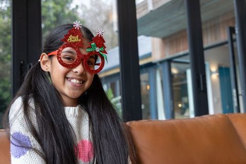 Indian girl wearing glitter christmas tree novelty glasses smiling on leather sofa